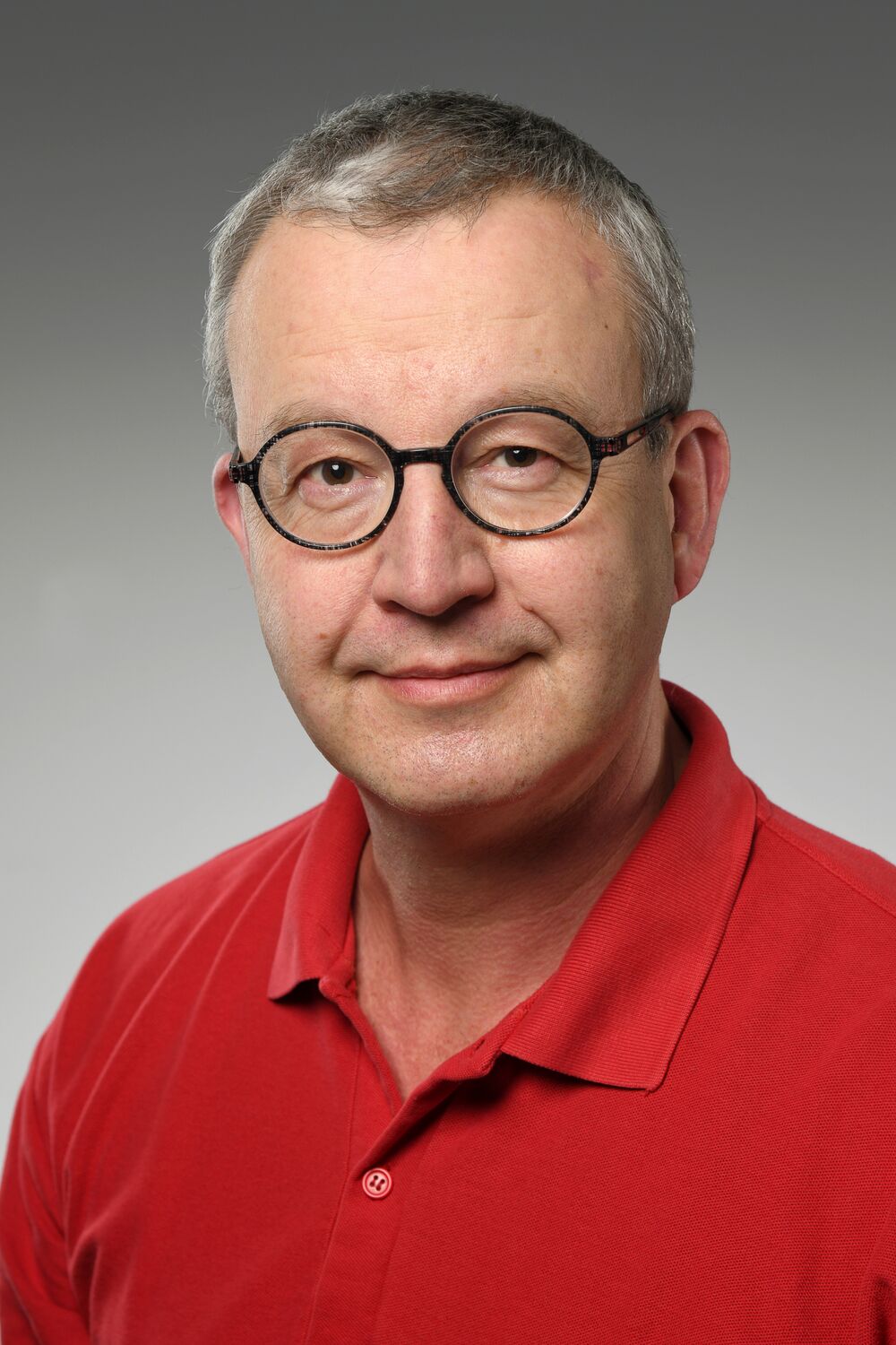 Portrait Dr. Belusa: Mann mittleren Alters mit kurzen Haaren und Brille mit runden Gläsern. Er trägt ein rotes Polo-Hemd und lächelt in die Kamera.